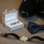 Accessoires d'un marié : alliances bicolores dans un écrin gris, nœud papillon à motifs, montre à bracelet noir et chaussures de ville en cuir, posés sur une surface texturée.